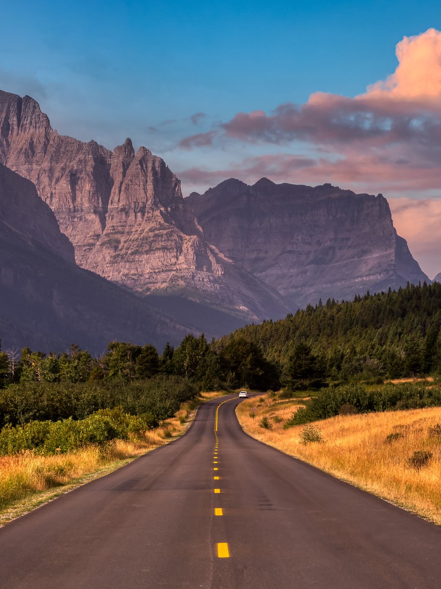 A two-lane highway running toward Rocky Mountain peaks under a colorful sunrise sky near St. Mary, Montana